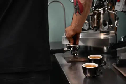 Barista z tatuażem na ramieniu przygotowuje kawę americana. Dwie filiżanki czarnej kawy stoją na blacie obok ekspresu.