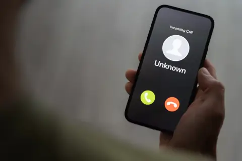 Dłoń trzyma smartfon z ekranem blokady, na którym widnieje "Incoming Call" od nieznanego numeru. Uważaj na vishing!