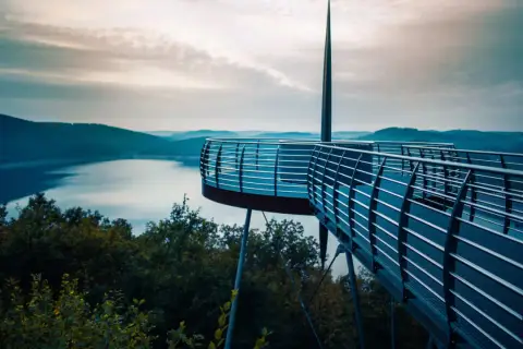 Moderne Aussichtsplattform über einem See, ideal für das Wandern in NRW. Sanfte Hügel und ein dramatischer Himmel im Hintergrund.