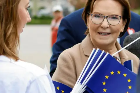Ewa Kopacz, gdzie mieszka, uśmiecha się, trzymając flagi UE.