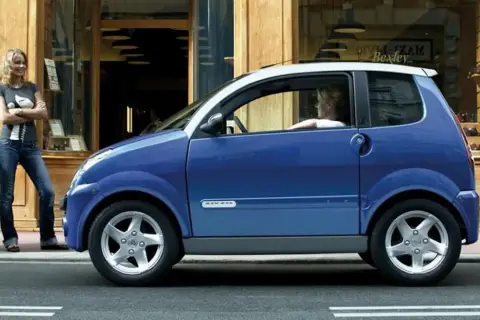 Niebieskie auto od 14 lat, z kobietą za kierownicą, stoi przed sklepem. Obok niego stoi druga kobieta.