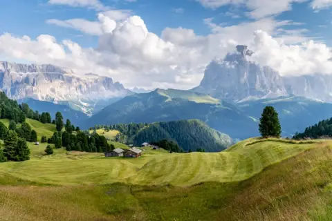 Zielone wzgórza i drewniane chatki w Val Gardena, z majestatycznymi Dolomitami w tle.