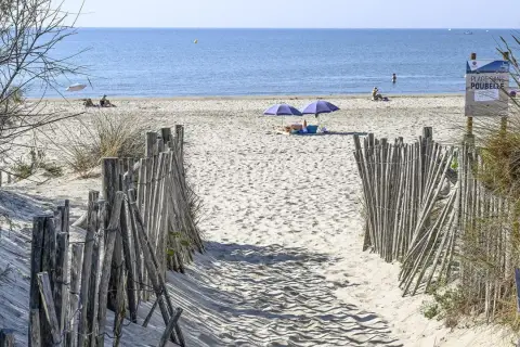 Ein sandiger Weg führt zum Montpellier Strand. Zwei Sonnenschirme spenden Schatten, während Menschen die Sonne genießen.