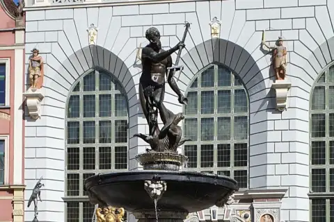 Fontanna Neptuna znajduje się w Gdańsku, przed fasadą budynku z arkadowymi oknami. Rzeźba przedstawia boga mórz na tle kamiennej architektury.