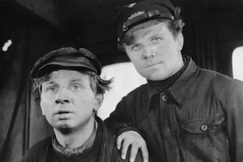Dwóch młodych mężczyzn w czapkach, w strojach roboczych, widać ich twarze. Jeden z nich opiera rękę na ramieniu drugiego. Scena z filmu człowiek na torze.