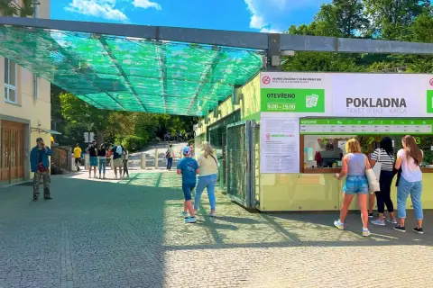 Wejście do ogrodu zoologicznego w Pradze. Ludzie stoją w kolejce do kasy biletowej pod zielonym zadaszeniem.