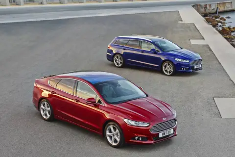 Dwa samochody Ford MK4: czerwony sedan i niebieskie kombi, stoją na asfaltowej nawierzchni przy betonowej konstrukcji i morzu.