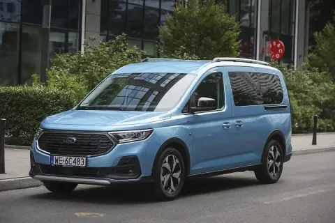 Błękitny ford 7 osobowy na tle nowoczesnego budynku. Idealny dla rodziny na wakacyjne wyprawy.