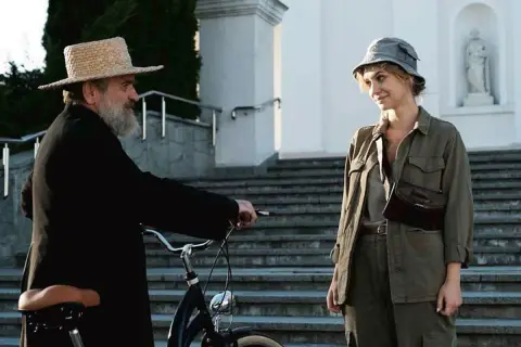 Scena z filmu Jacka Bromskiego: mężczyzna w słomkowym kapeluszu i czarnej sutannie przy rowerze rozmawia z kobietą w wojskowym kapeluszu.