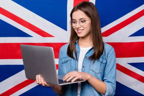 Uśmiechnięta dziewczyna w okularach pracuje na laptopie na tle flagi Wielkiej Brytanii, tworząc przykładowy artykuł po angielsku.