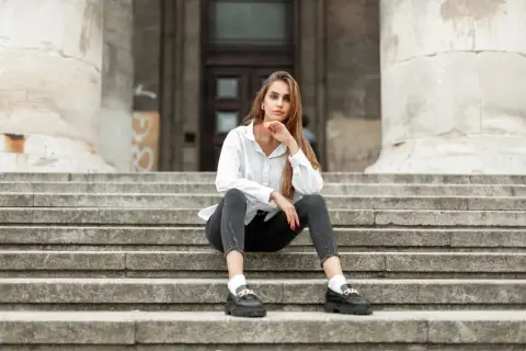 Eleganckie mokasyny to idealne buty do szarych spodni. Kobieta w białej koszuli i czarnych jeansach siedzi na schodach.