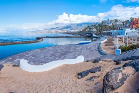 Słoneczna playa de las americas z basenem, piaskiem i kolorowymi budynkami w tle.