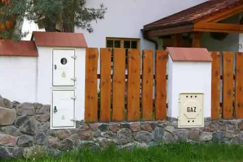 Zastanawiasz się, ile kosztuje przyłącze gazu? Na zdjęciu widzimy skrzynkę z napisem "GAZ" obok drewnianego płotu i kamiennego murku.