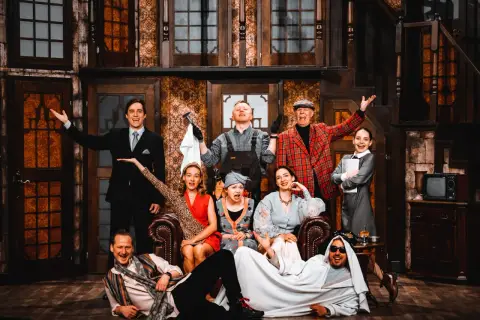 Aktorzy w teatralnej scenografii, jakby w teatrze bez rzędów, pozują do zdjęcia. Wszyscy uśmiechnięci, w barwnych strojach, tworzą radosną atmosferę.