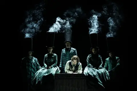 Scen från Sweeney Todd operan. Människor med höga hattar och rök som stiger ur dem. En man skriver vid ett bord.