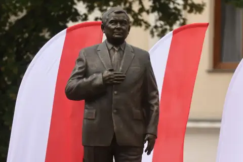 Pomnik Lecha Kaczyńskiego, jednego z wielu w Polsce, stoi na tle biało-czerwonych flag.