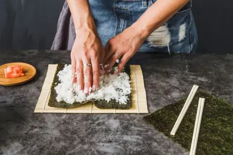Dłonie rozprowadzają ryż na nori, przygotowując zaprawę do sushi na 500g ryżu. Obok leżą pałeczki i marynowany imbir.