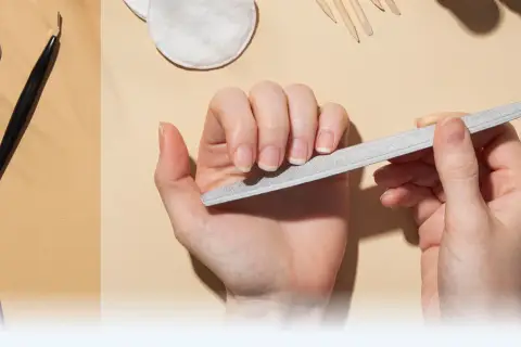Dłonie pielęgnują paznokcie, używając pilnika. W tle widać narzędzia do manicure, takie jak nożyczki i cążki, tworząc kompletny zestaw do pielęgnacji macierzy paznokcia.