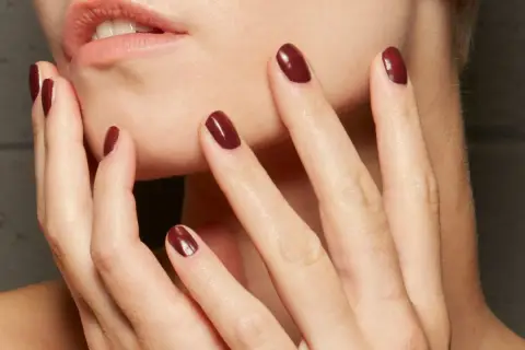 Dłonie z idealnym, ciemnoczerwonym manicurem kombinowanym. Delikatne muśnięcie policzka podkreśla elegancję.