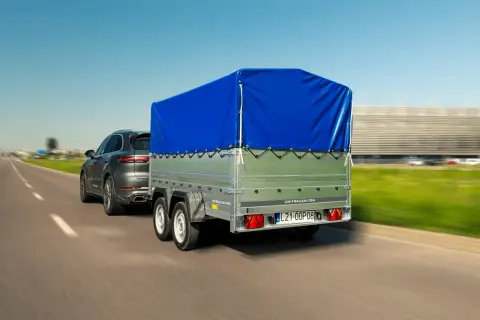 Przyczepa kempingowa bez prawa rejestracji, z niebieską plandeką, ciągnięta przez ciemny SUV.