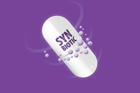 Biała kapsułka z napisem "SYN BIOTIC" na fioletowym tle. Drobne kuleczki unoszą się wokół. Dowiedz się, synbiotyk co to.