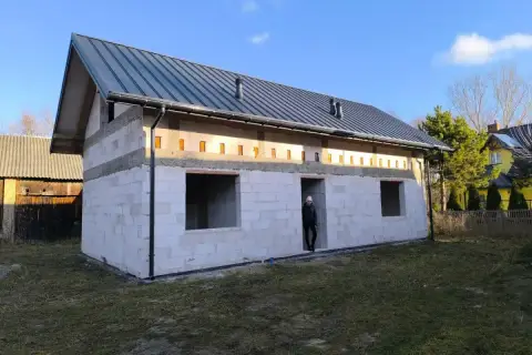 Budowa domu bez pozwolenia w toku. Białe bloczki, szary dach, puste okna. Ktoś stoi w drzwiach.