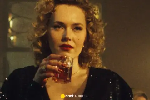 Kobieta z burzą blond loków i czerwoną szminką, pijąca drinka. W tle widać fragmenty scenografii, sugerujące klimat filmu "Jak pokochałam gangstera obsada".