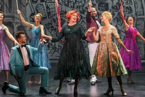 Scen från "Tootsie" på Oscarsteatern. En man i ljusblå kostym knäböjer medan en kvinna i glittrig grön klänning och andra i färgglada klänningar dansar.
