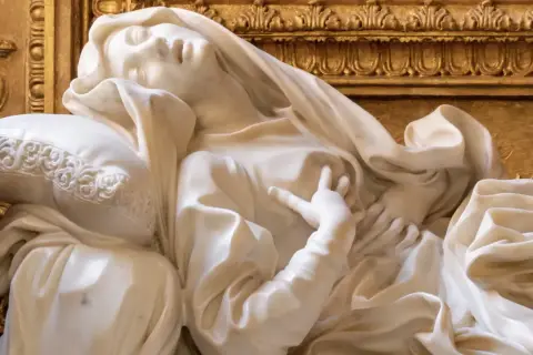 Estasi di Santa Teresa a Roma, marmo bianco su fondo dorato. La santa giace su un cuscino, con un'espressione di profonda emozione.
