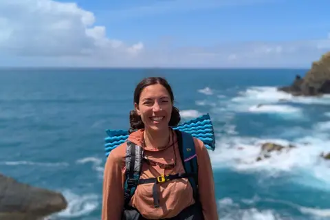 A smiling hiker with a backpack and sleeping pad enjoys the view of the ocean waves crashing on the shore along the Fischerweg trail in Portugal.