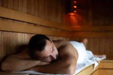 Mężczyzna odpoczywa w saunie po treningu, otulony białym ręcznikiem.