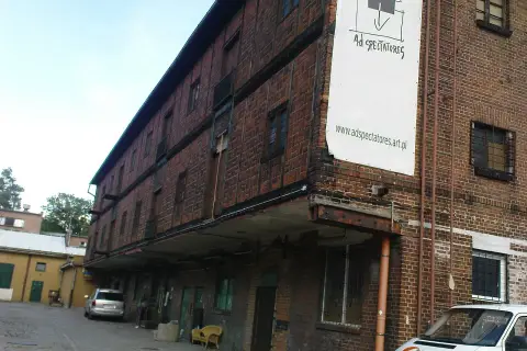 Stara ceglana kamienica z banerem "Ad Spectatores". Obok samochody i żółte krzesła.