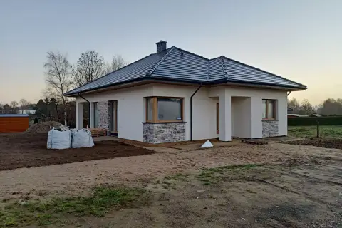 Nowy, nowoczesny dom w renklodach 15 z szarym dachem i białymi ścianami, otoczony placem budowy.