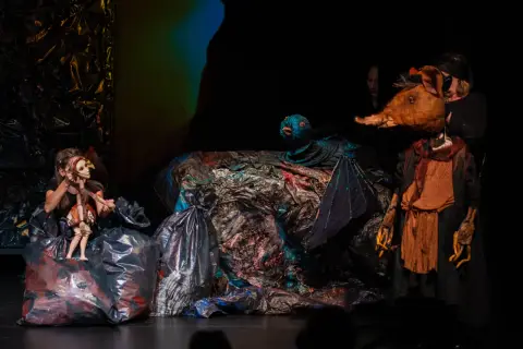 Na scenie teatru lalki i aktora w Łomży, dziewczynka animuje drewnianą lalkę, obok niej ptak i postać z głową zwierzęcia.