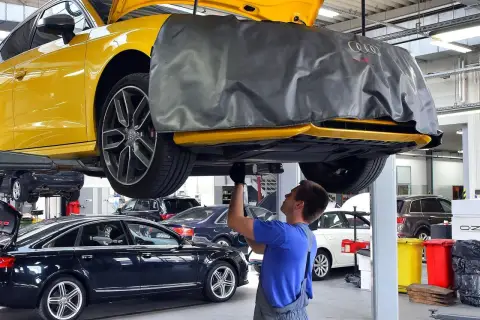 Mechanik pracuje pod żółtym Audi, sprawdzając, ile kosztuje wymiana oleju silnikowego w samochodzie. W tle inne auta.