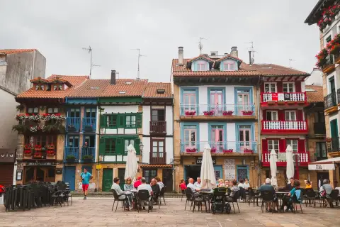 Fachada de casas coloridas en Hondarribi, con balcones floridos y gente disfrutando en terrazas.