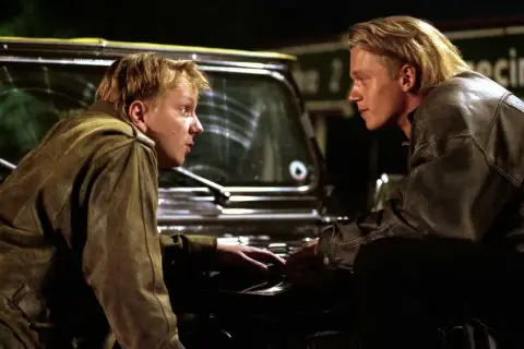 Dwóch młodych mężczyzn w skórzanych kurtkach, jak z filmów o mafii, pochyla się nad maską samochodu, prowadząc intensywną rozmowę.