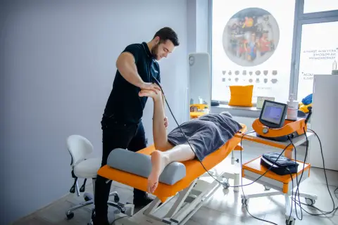 Fizjoterapeuta wykonuje zabieg falą uderzeniową na stopę pacjenta.