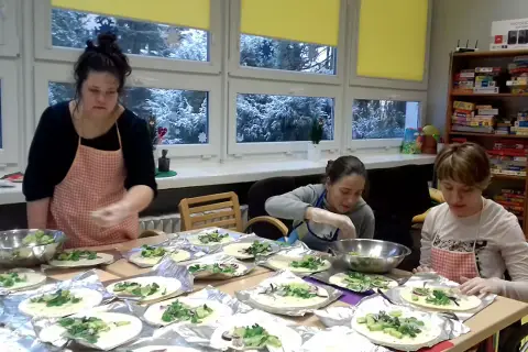 Trzy kobiety w fartuchach przygotowują jedzenie na stole, na tle okien ze śniegiem.