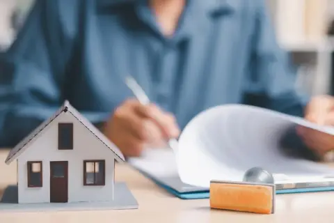 Model domu i pieczątka obok dokumentów, na których ktoś pisze. To symbol finalizacji umowy deweloperskiej.