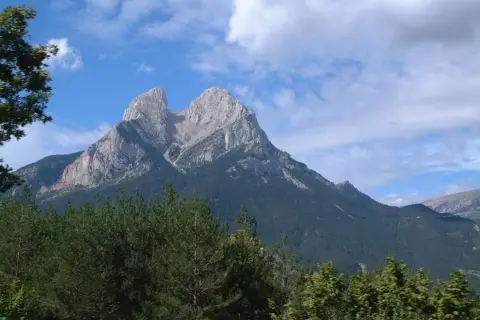 Majestuoso Pedraforca se alza imponente, su altitud desafía las nubes. Bosques verdes cubren sus faldas bajo un cielo azul.