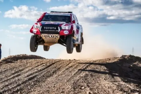 Toyota Hilux w locie podczas rajdu Dakar. Widzowie obserwują zmagania na pustynnym terenie.