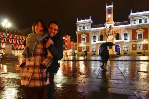 Pareja sonriente en la Plaza Mayor de Valladolid, un lugar bonito que ver en Valladolid, con edificios iluminados y el suelo mojado por la lluvia.