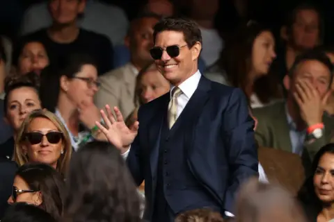 Tom Cruise w garniturze, w okularach przeciwsłonecznych, macha do publiczności. Zastanawiasz się, ile zarabia Tom Cruise?