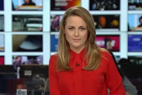 Marta Kłos, której wiek nie jest widoczny, w czerwonej bluzce z kokardą, na tle ekranów informacyjnych.