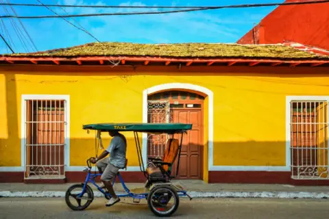 Un hombre pedalea un taxi bicitaxi frente a una casa amarilla vibrante, un recuerdo de mi viaje a Cuba.