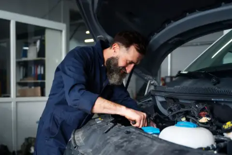 Mechanik z brodą pracuje przy silniku. Zastanawiasz się, ile kosztuje wymiana chłodnicy w samochodzie?
