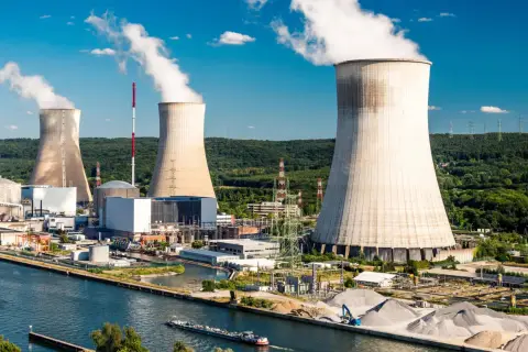 Ogromne chłodnie kominowe elektrowni atomowej wyrzucają białą parę na tle błękitnego nieba. Woda w kanale odbija słońce.