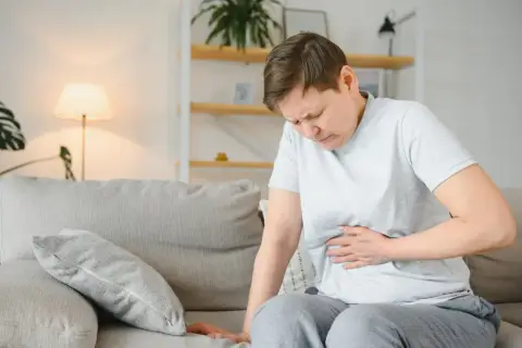 Kobieta siedzi na kanapie, trzymając się za brzuch. Jej twarz wyraża ból, co może sugerować przewlekłe zapalenie trzustki.