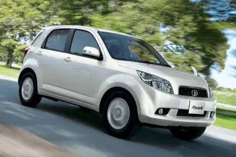 Biały Toyota Rush pędzi po drodze, otoczony zielenią drzew.